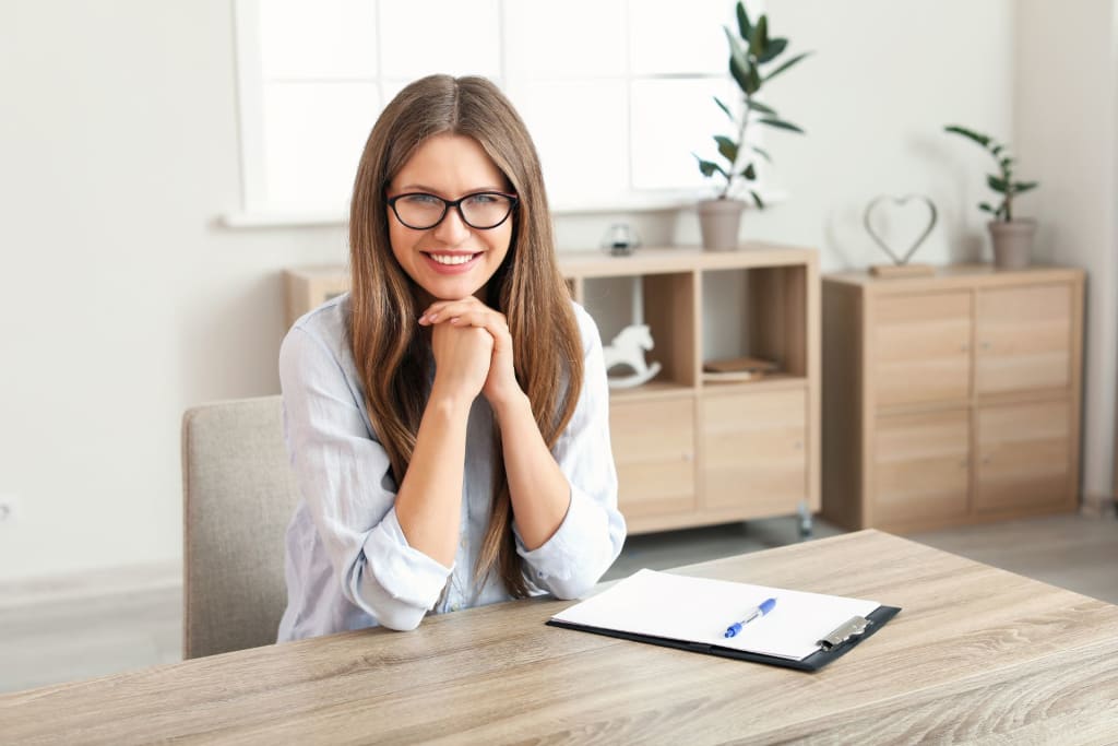 A woman with long brown hair and glasses sits at a desk, smiling, with a clipboard and pen in front of her—an inviting space for a private practice consultant; the background includes shelves and potted plants.